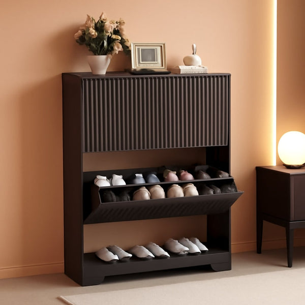 Black fluted shoe cabinet with the flip drawer open showing shoes stored at an angle, and an open shelf below, in a warm lit entryway
