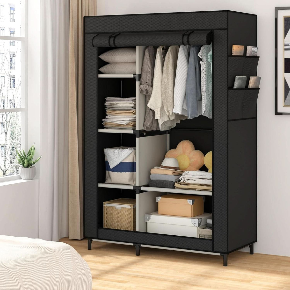 Black portable closet in a bright bedroom beside a window, holding hanging shirts, folded towels and storage boxes