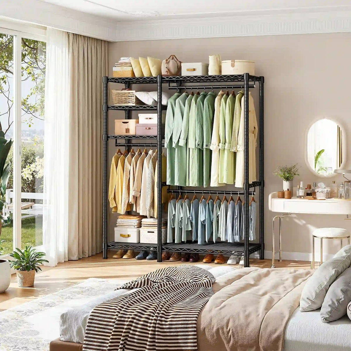 Black rolling clothes rack against a bedroom wall beside a window, holding hanging shirts, folded stacks and storage boxes