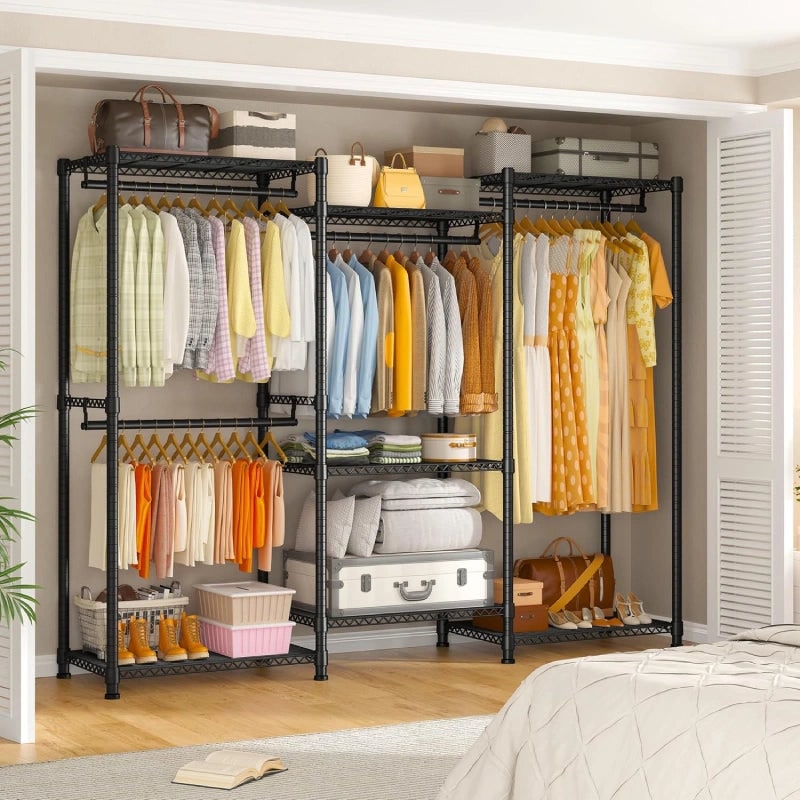 Black freestanding closet rack along a bedroom wall next to a bed, filled with hanging clothes, folded linens and storage boxes