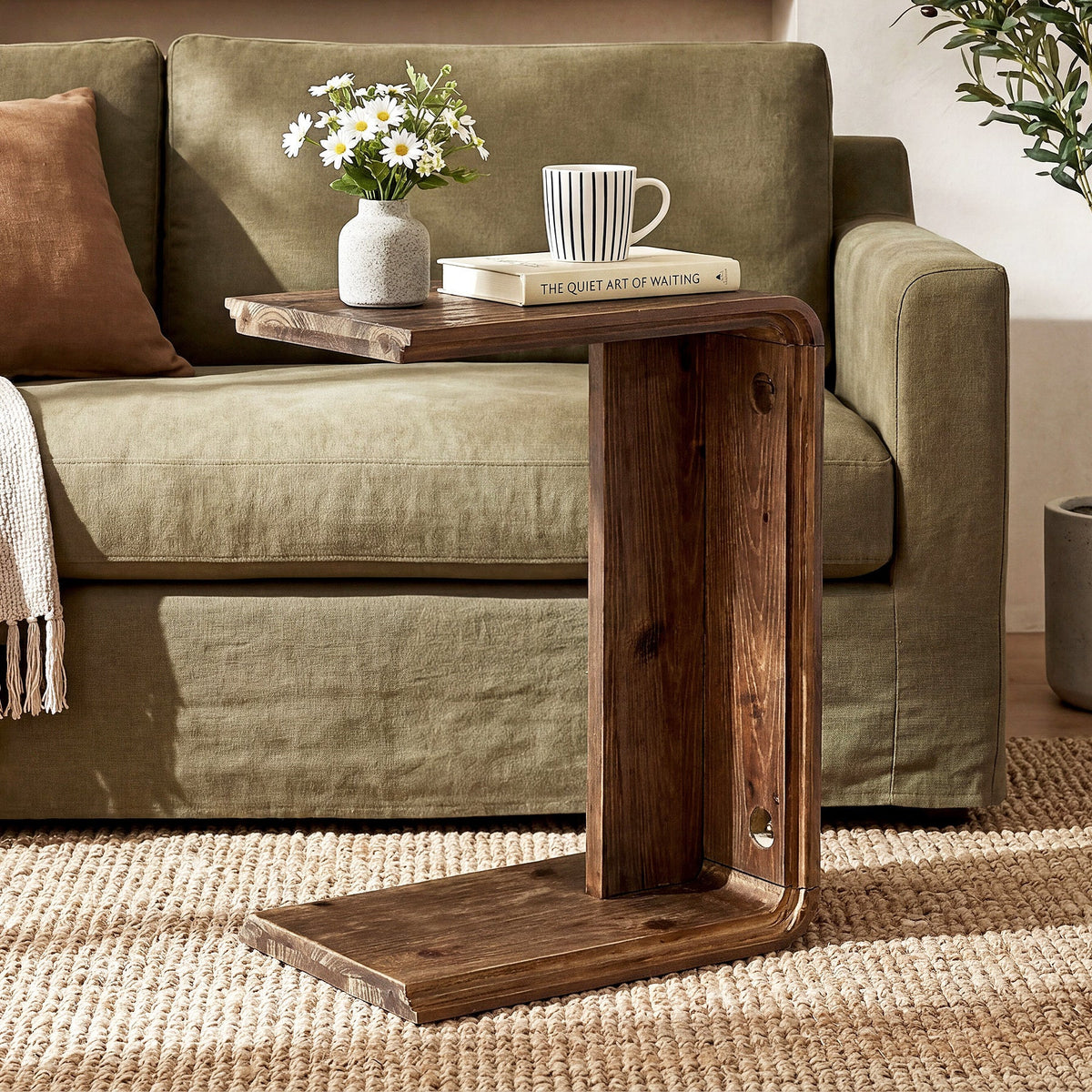 Natural wood C table tucked over a sofa arm cushion with flowers and a mug