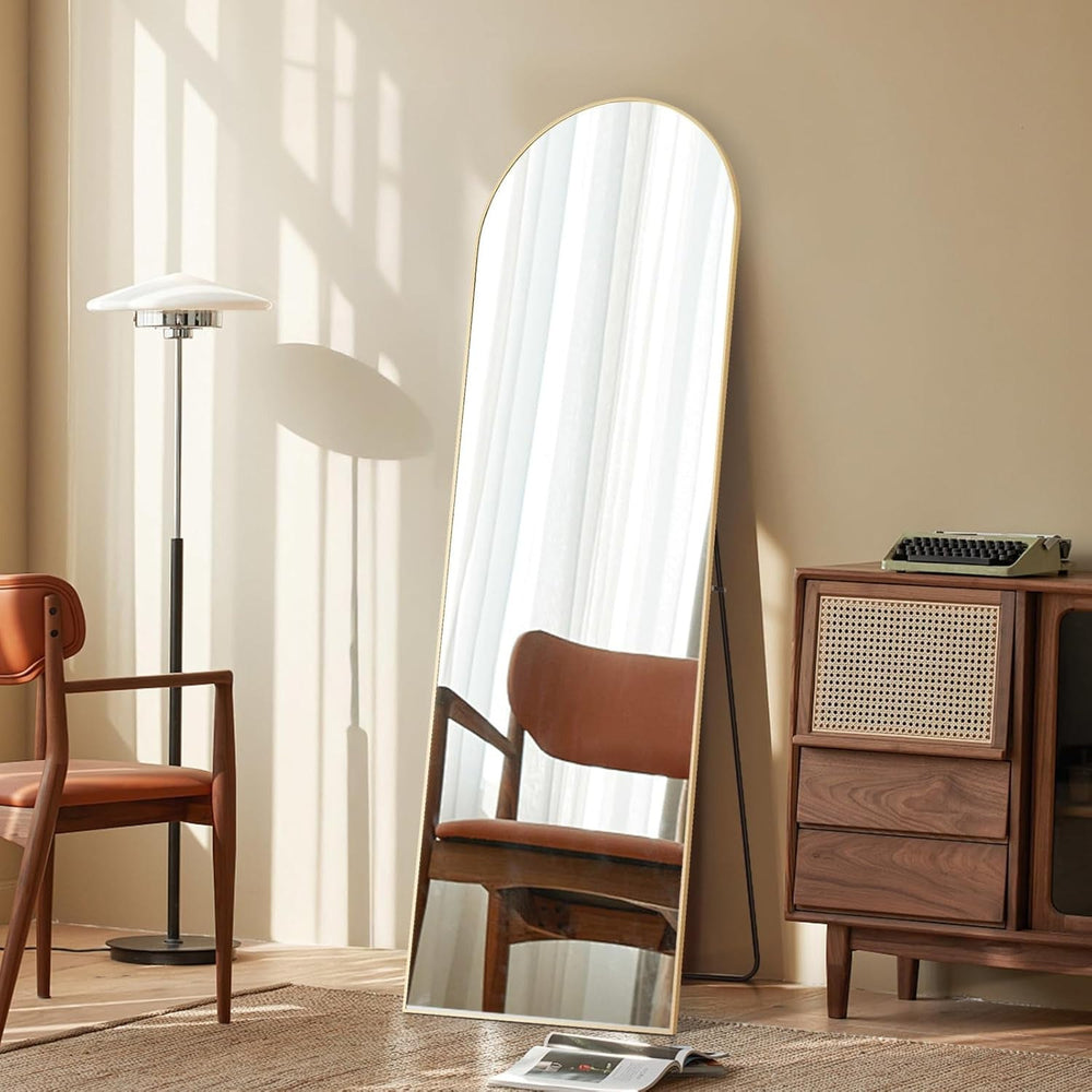 Stowena 64 inch arched full length mirror in gold leaning by a reading chair
