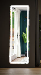 Rounded-corner LED full length mirror hung on a dark wall, reflecting a window, curtain and plant