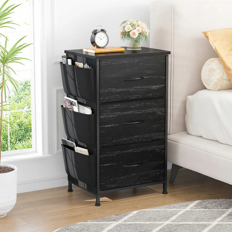 Stowena black 3 drawer nightstand with side pockets beside a bed with white bedding
