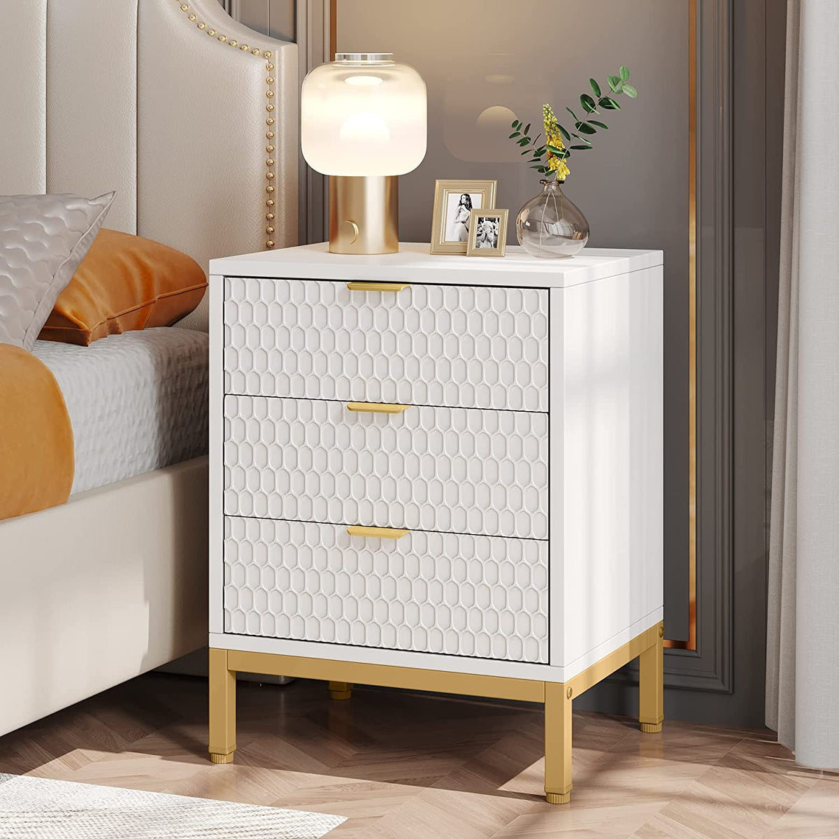 Stowena white and gold 3 drawer nightstand beside an upholstered bed with a glowing lamp